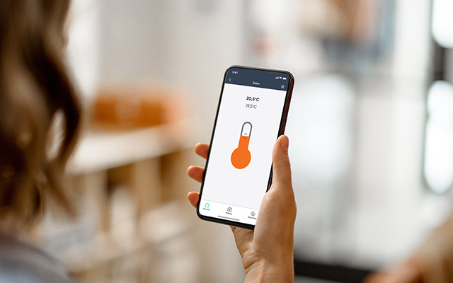 Un visuel d'une femme regardant son téléphone. Sur l'écran on peut voir l'application, mettant en avant la programmation chauffage.